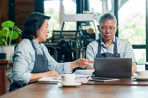 Senior asian couple cafe owners analyzing data and making decision using laptop in restaurant, showing cooperation and partnership in managing small family business successfully